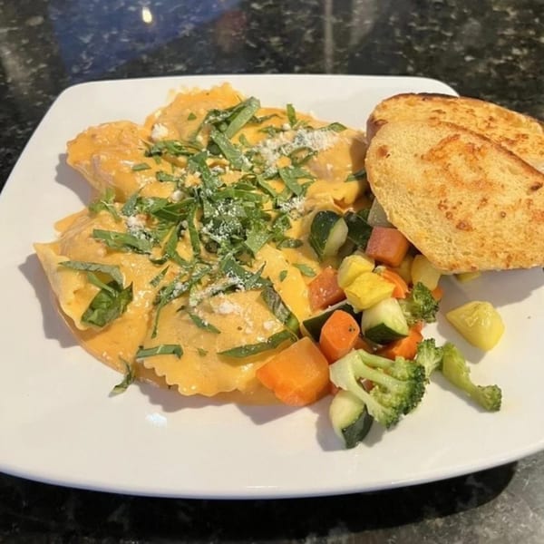 Ravioli with creamy sauce, fresh herbs, mixed vegetables, and toasted bread on a white plate.