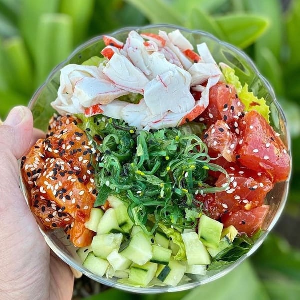 A vibrant poke bowl with crab, tuna, salmon, seaweed, and cucumber, held against a green background.