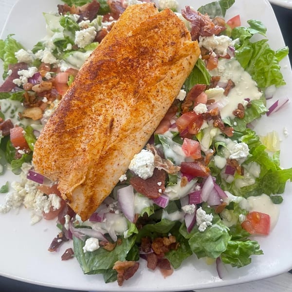 Seared fish fillet on a bed of green salad with bacon, tomatoes, red onion, and crumbled cheese.