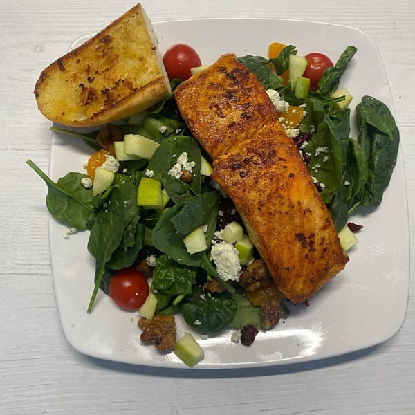 Grilled salmon fillet on a spinach salad with tomatoes, apples, and feta, served with garlic bread.